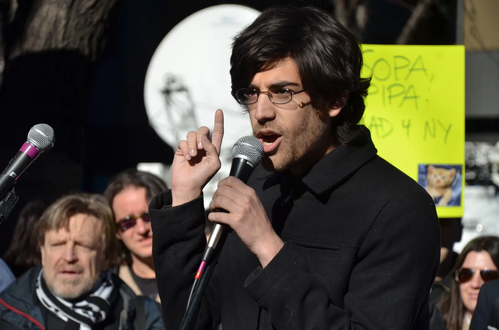 Aaron Swartz speaking into a microphone at an outdoor rally, one finger raised. Behind him a hand-lettered yellow sign reads SOPA, PIPA: BAD 4 NY.
